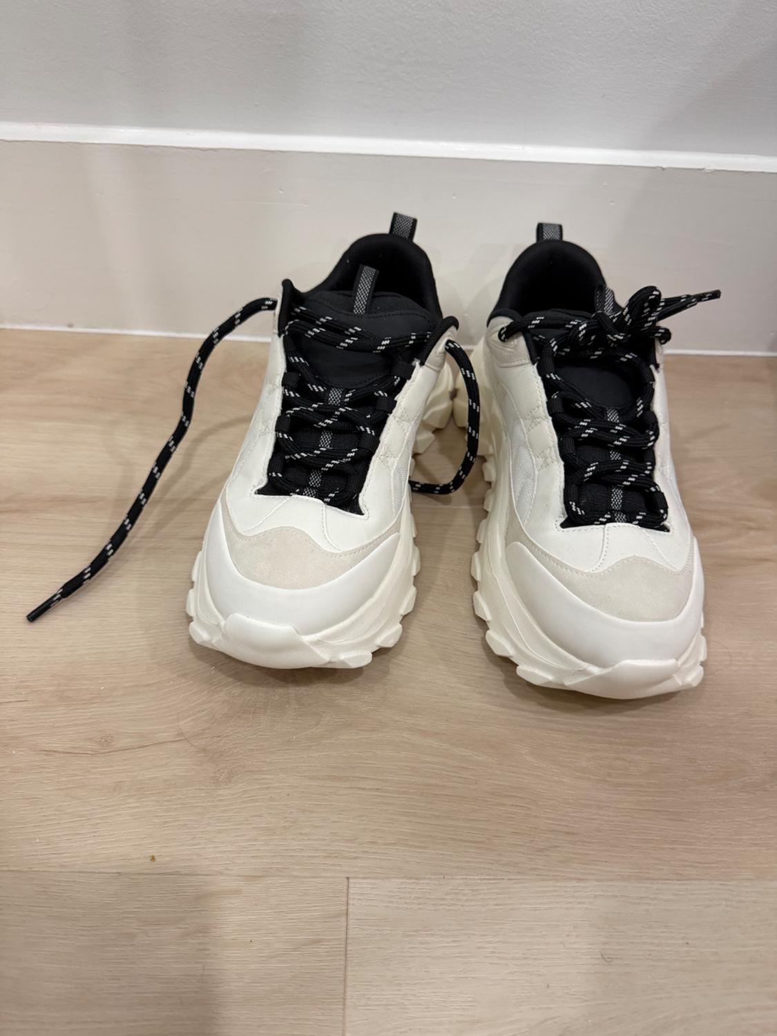 A pair of white athletic sneakers with a thick sole and black laces with white details, on a light wooden floor.