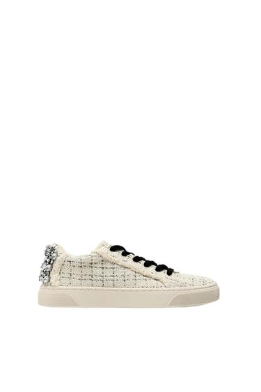 Beige trainer with woven texture, black laces and jewelled detail on the heel, on a white background.