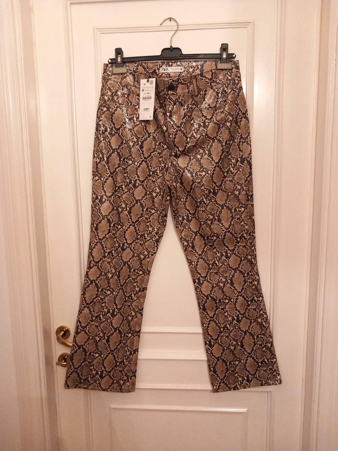 Snake-print trousers hanging on a hanger against a white door. ZARA text visible on the label.