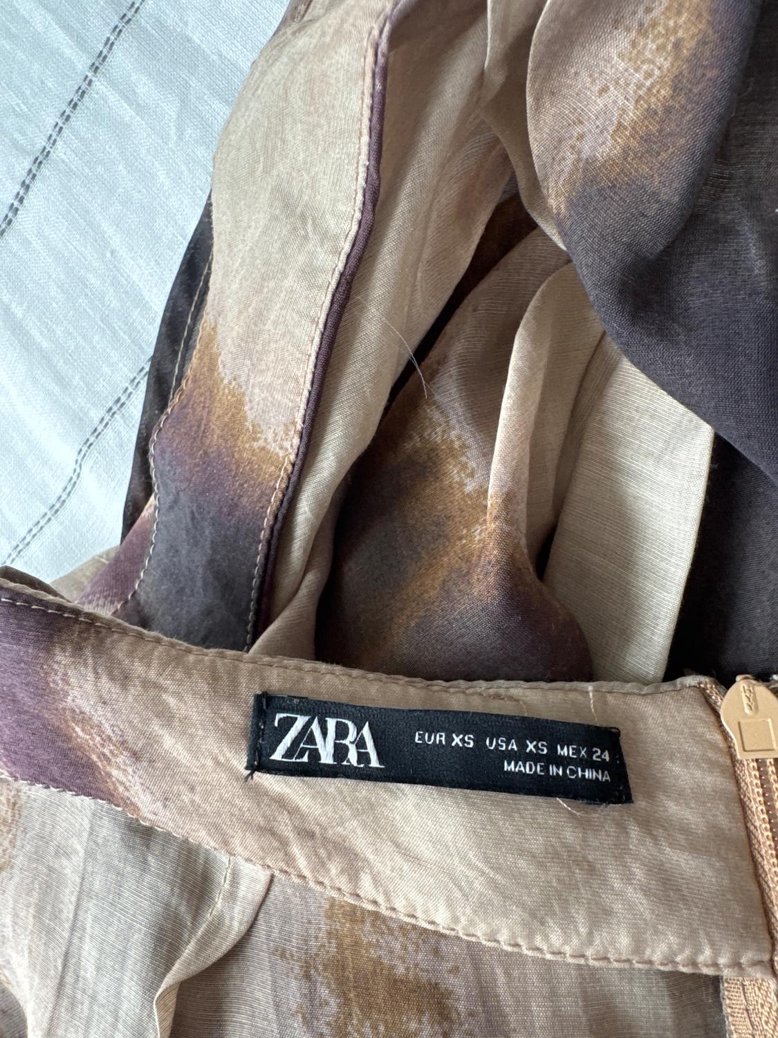 Zara clothing label in brown and beige fabric with a gold zip. Text: "EUR XS USA XS MEX 24 MADE IN CHINA".