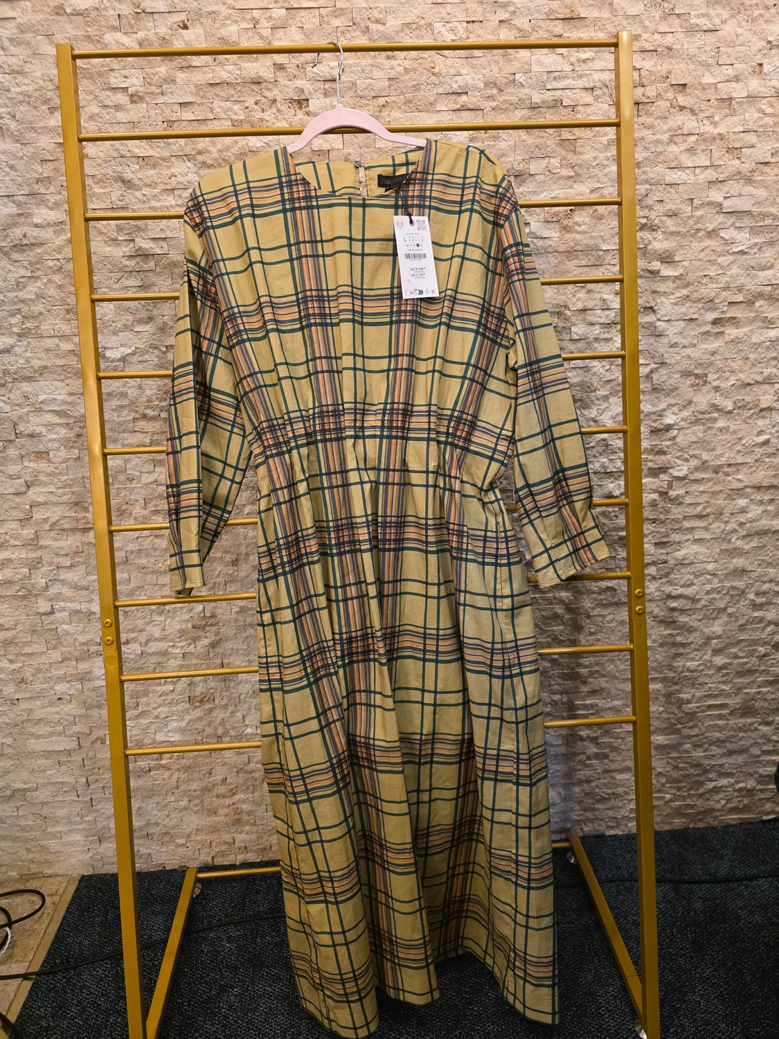 Yellow and green plaid dress hanging on a gold rack in front of a light stone wall. Label visible on the collar.
