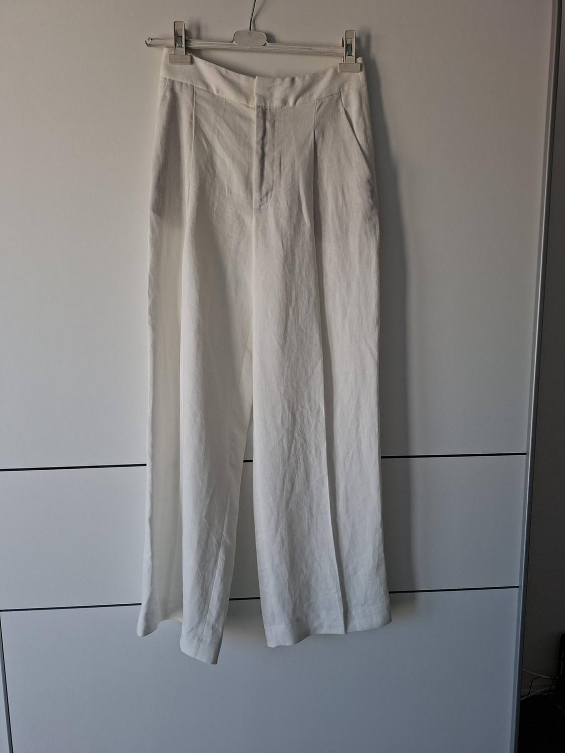 White wide-leg trousers with pleats, hanging from a hanger in front of a white background with black horizontal lines.