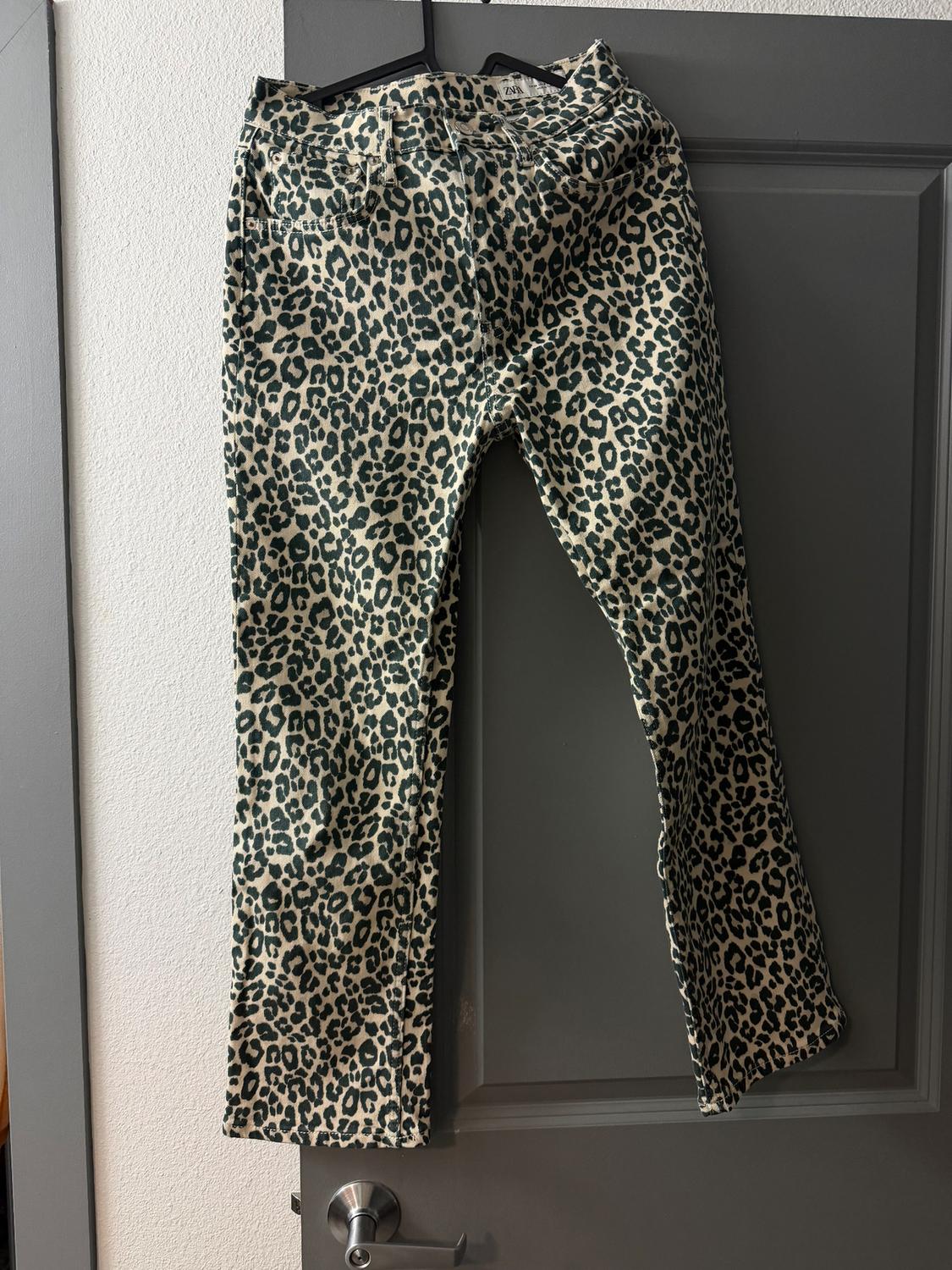 Leopard print trousers hanging on a grey door. White background and visible tag on the waistband.