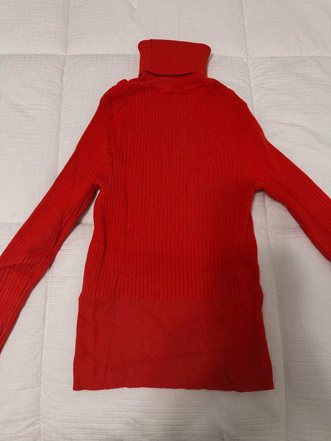 Red ribbed turtleneck sweater with long sleeves, on a white quilted bedspread. Casual style, relaxed vibe.