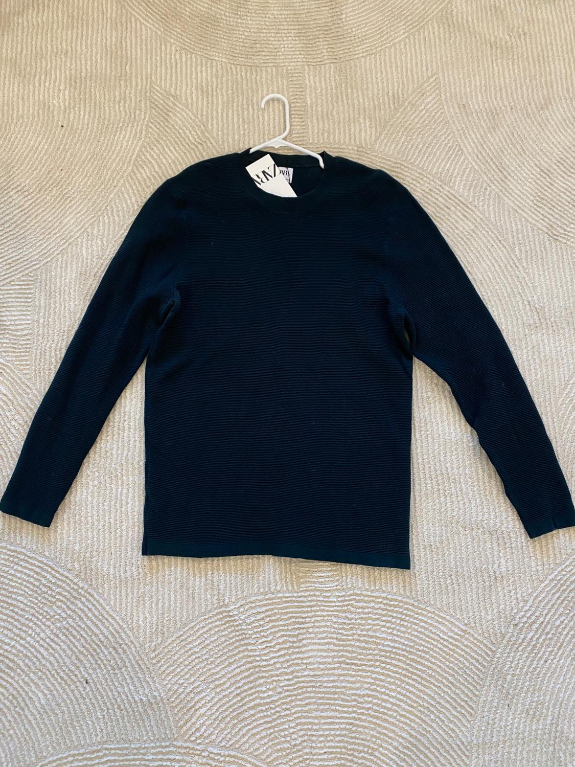 Black long-sleeved jumper on a beige rug with a circular design. Visible tags on the collar.