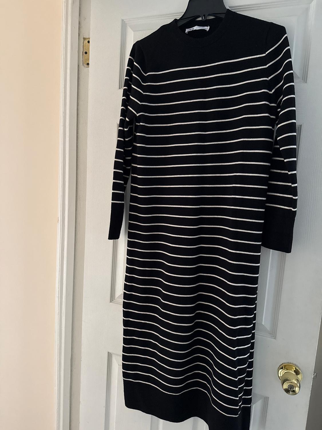 Black dress with white stripes hanging on a white door. Long sleeves and round neckline. Illuminated indoor environment.
