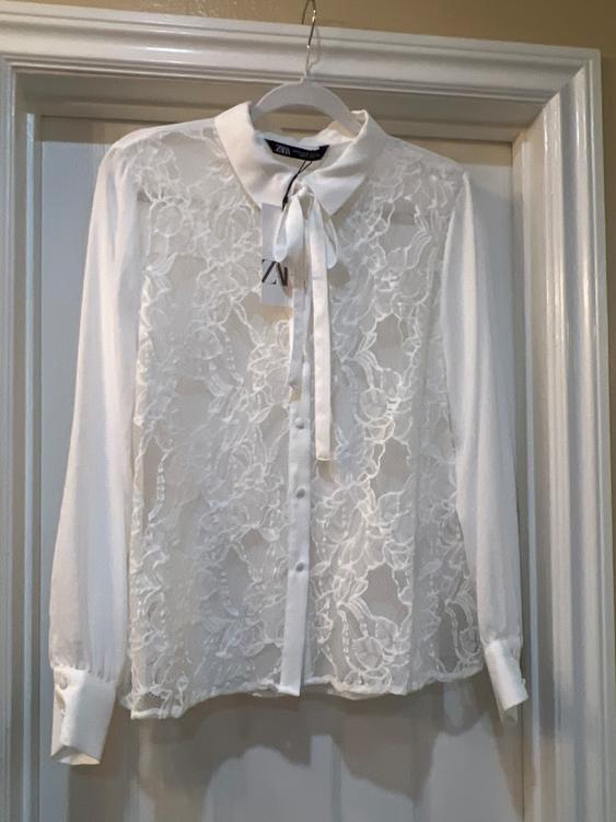 White lace blouse with a bow at the neck, hanging on a hanger. Visible tag with text "ZVN". White door background.