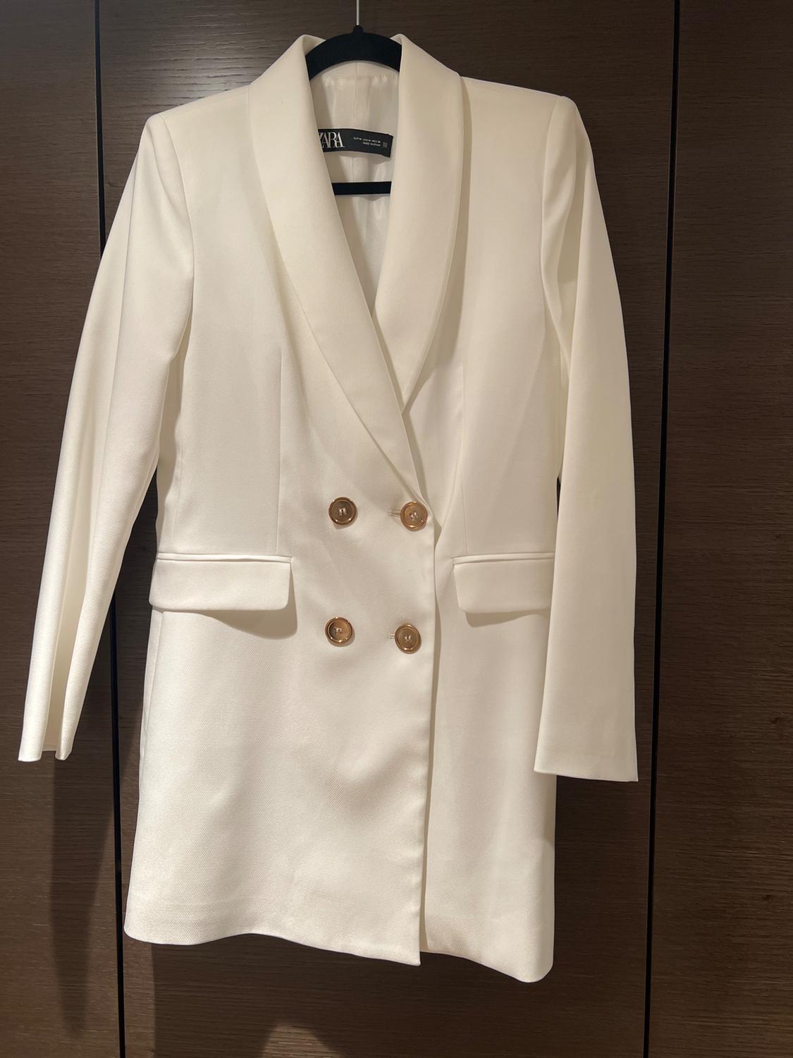 Double-breasted white blazer with gold buttons, hanging on a hanger against a dark wood background.