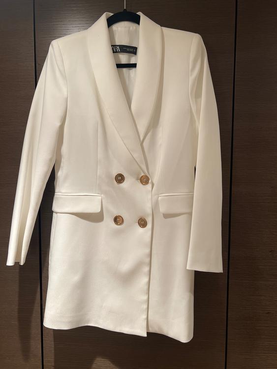 Double-breasted white blazer with gold buttons, hanging on a hanger against a dark wood background.