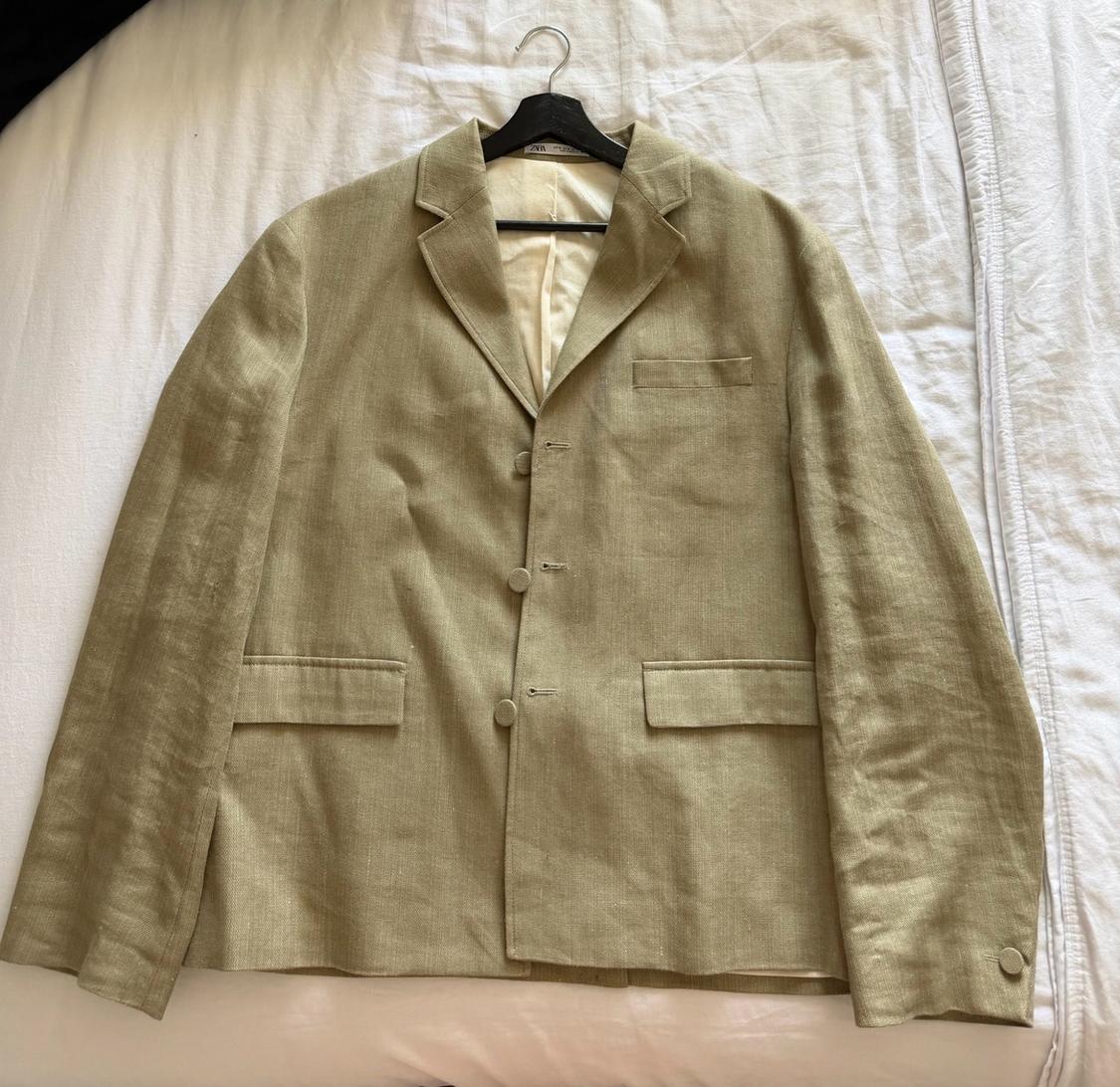Olive green three-button blazer with pockets, hanging on a black hanger over crumpled white sheets.