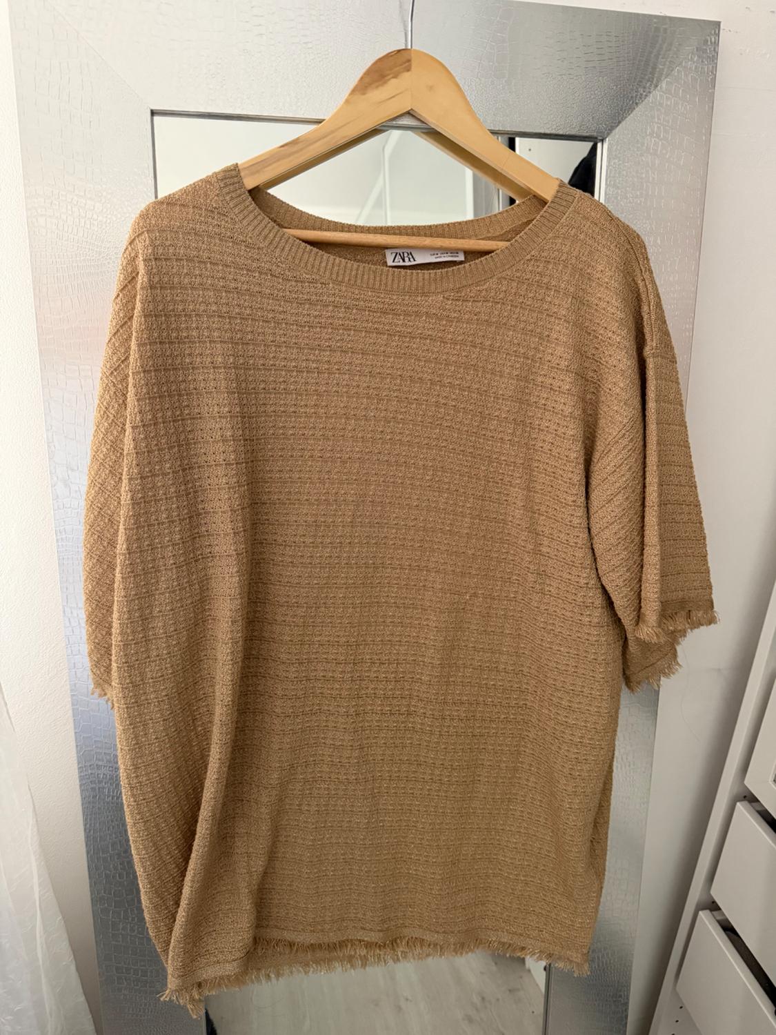 Beige textured fabric T-shirt hanging on a wooden hanger in front of a mirror. Visible label: "Zara".