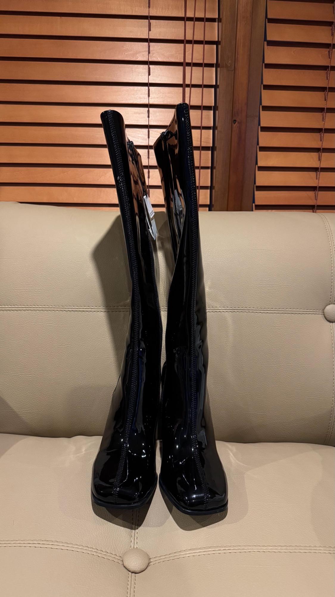 Shiny black square-toe high boots with a front zip on a beige sofa, with wooden blinds in the background.