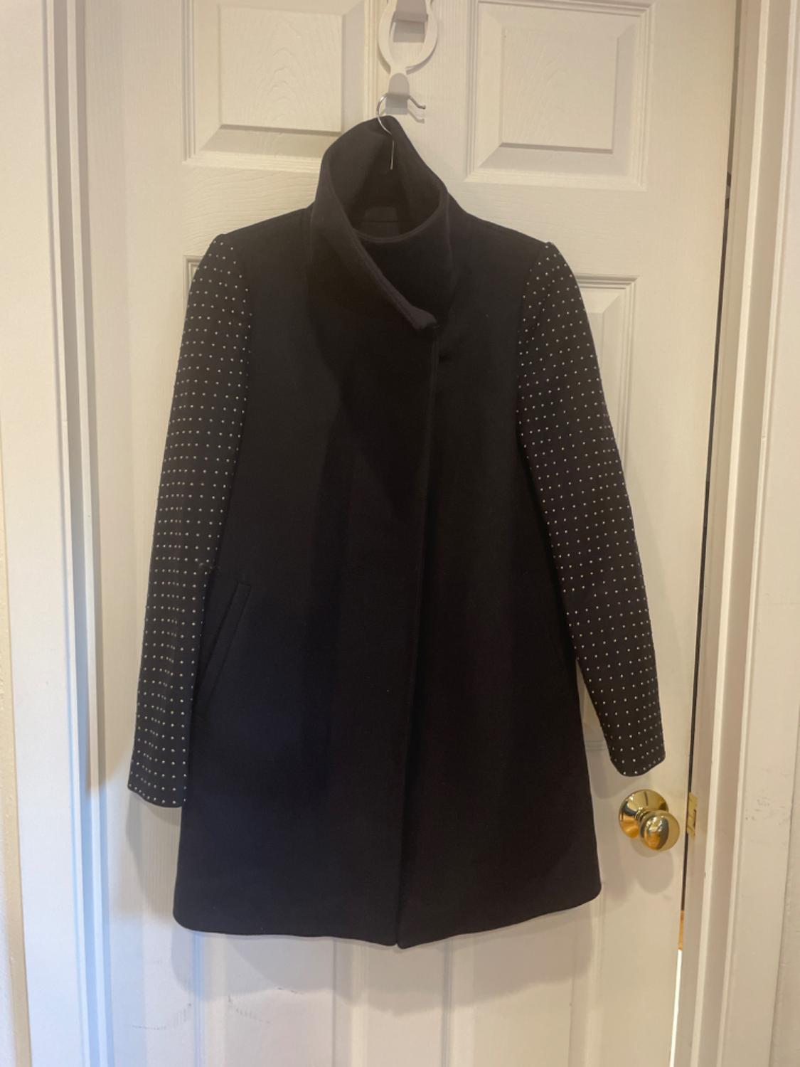 Front view of a black coat with a high neck and sleeves with white dots, side pockets.