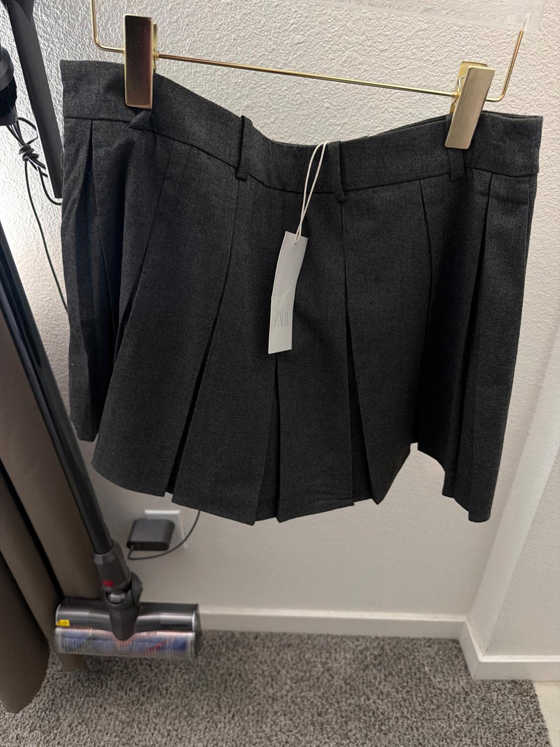 Gray pleated skirt hanging on a golden hanger against a white wall. Visible tag with the text "Zara". Carpeted floor.