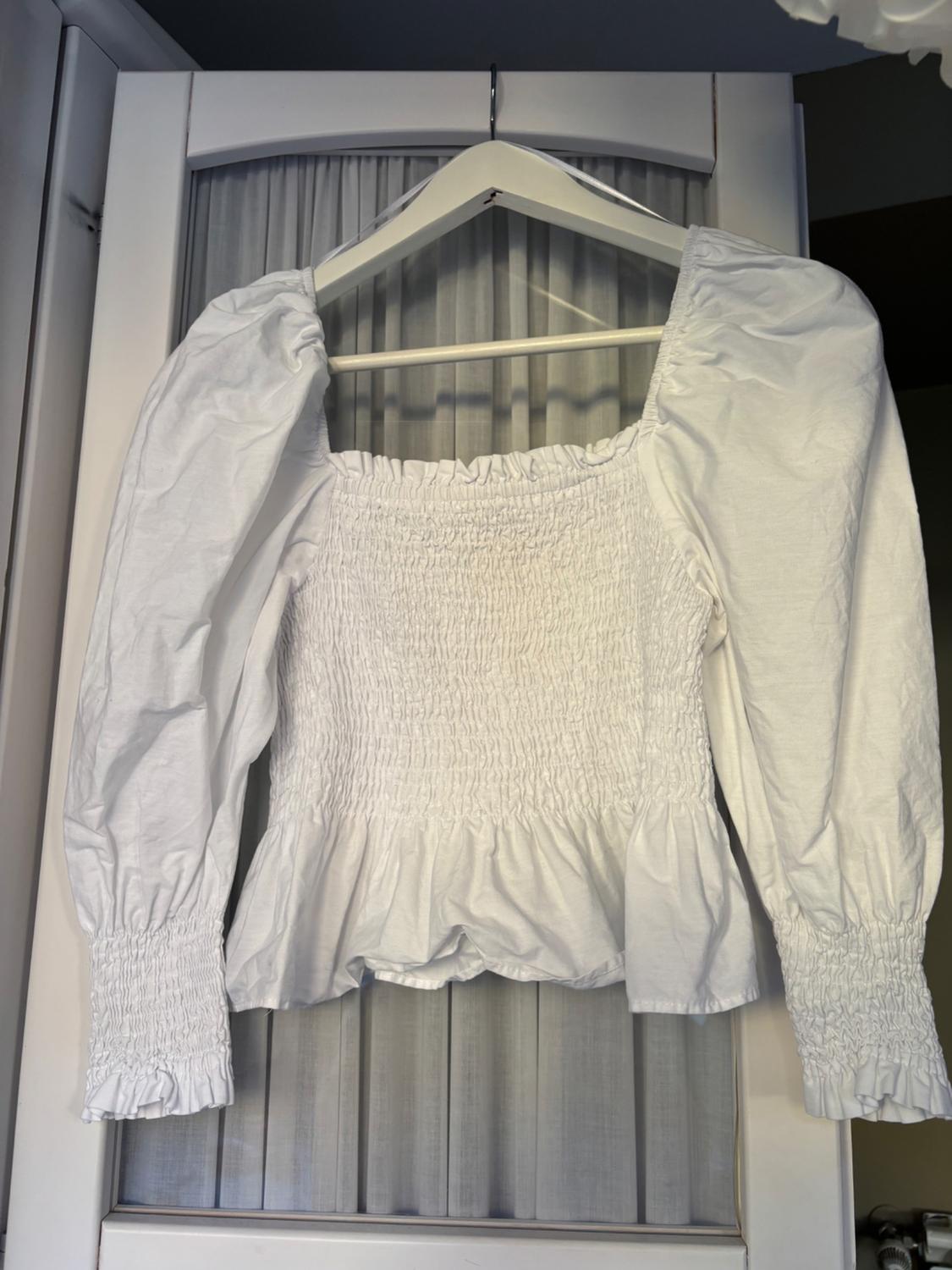 White, long-sleeved blouse with a ruched design and frilled trim, hanging on a hanger in front of a door with sheer curtains.