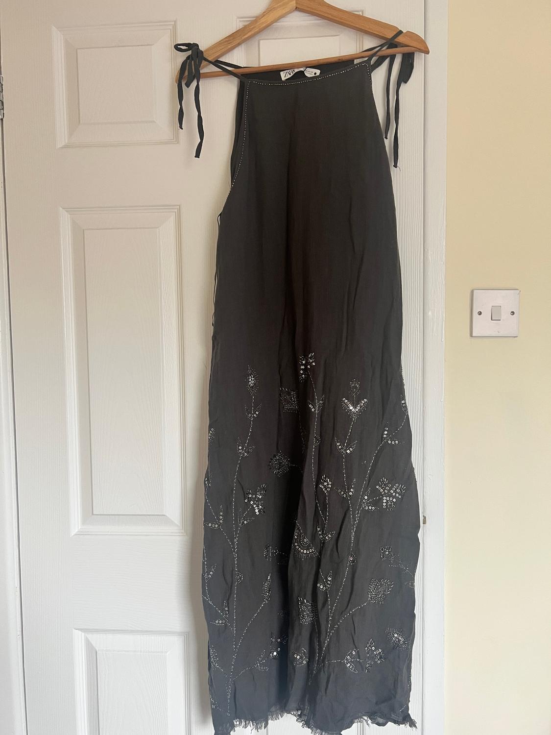 Black dress hanging on a wooden hanger, with embroidered floral details on the bottom. Background: white door.