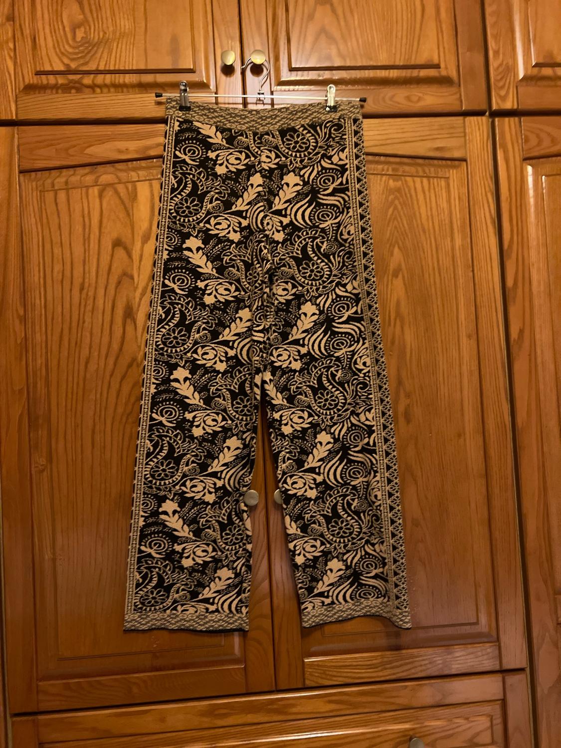 Black and beige floral print trousers, hanging on a hanger in front of a classic wooden wardrobe in a relaxed setting.