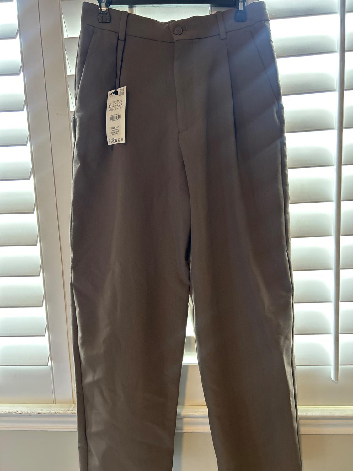 Brown pants hanging in front of a window with white blinds. Visible tag with product details. Natural light.