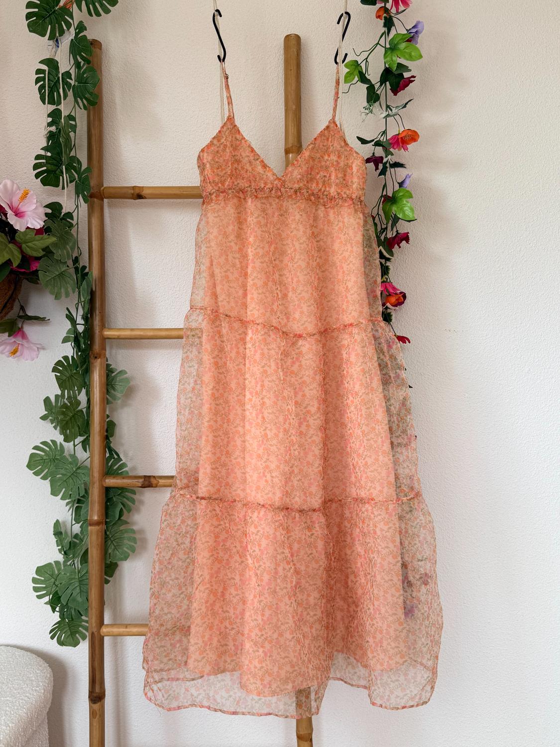 Long, peach-toned, spaghetti-strap dress with a floral print, hung on a bamboo ladder decorated with leaves and flowers.