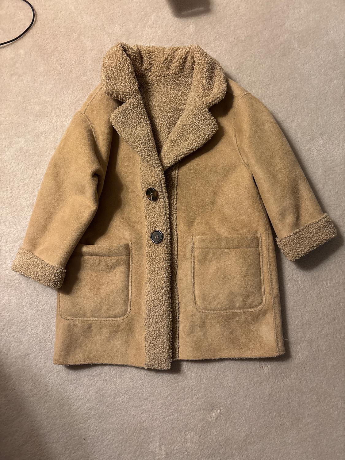 Light brown coat with fleece lining, two front pockets and button fastening, on a beige background.