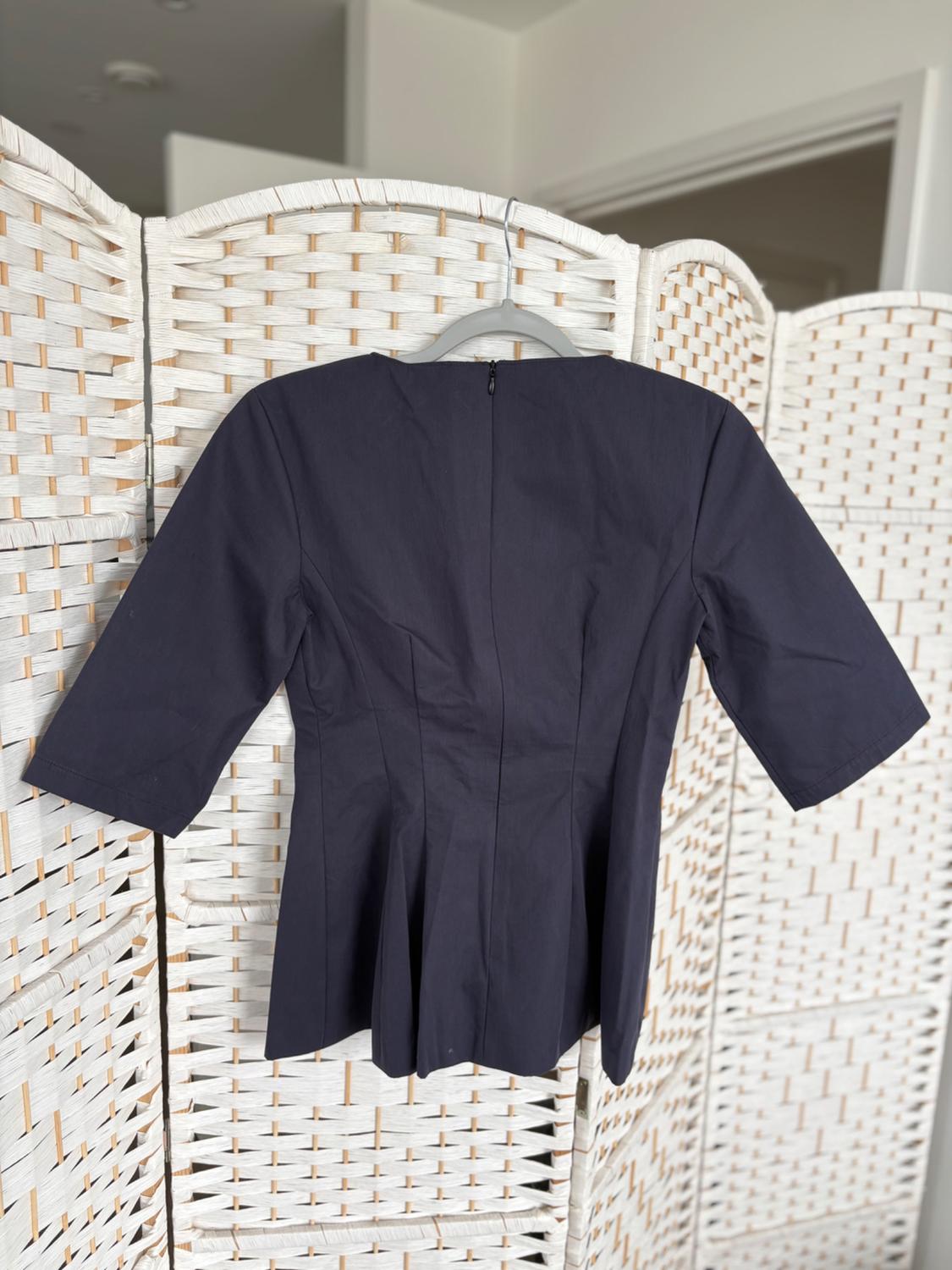 A black three-quarter sleeve blouse is hanging on a white woven screen. The setting is indoors and has natural light.