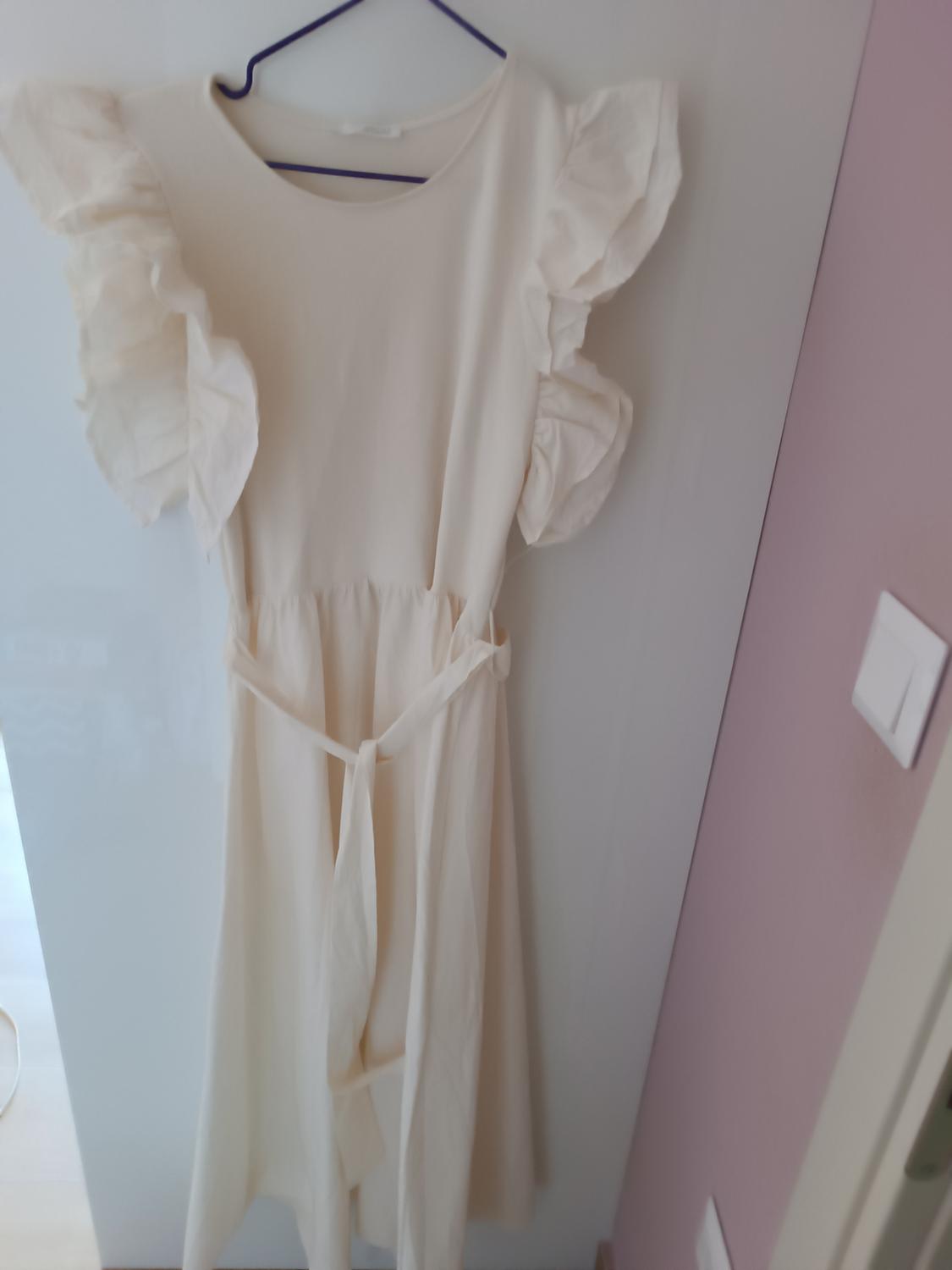 White dress with frills on the sleeves, hanging on a blue hanger over a white door. Background with pink wall and visible plug socket.