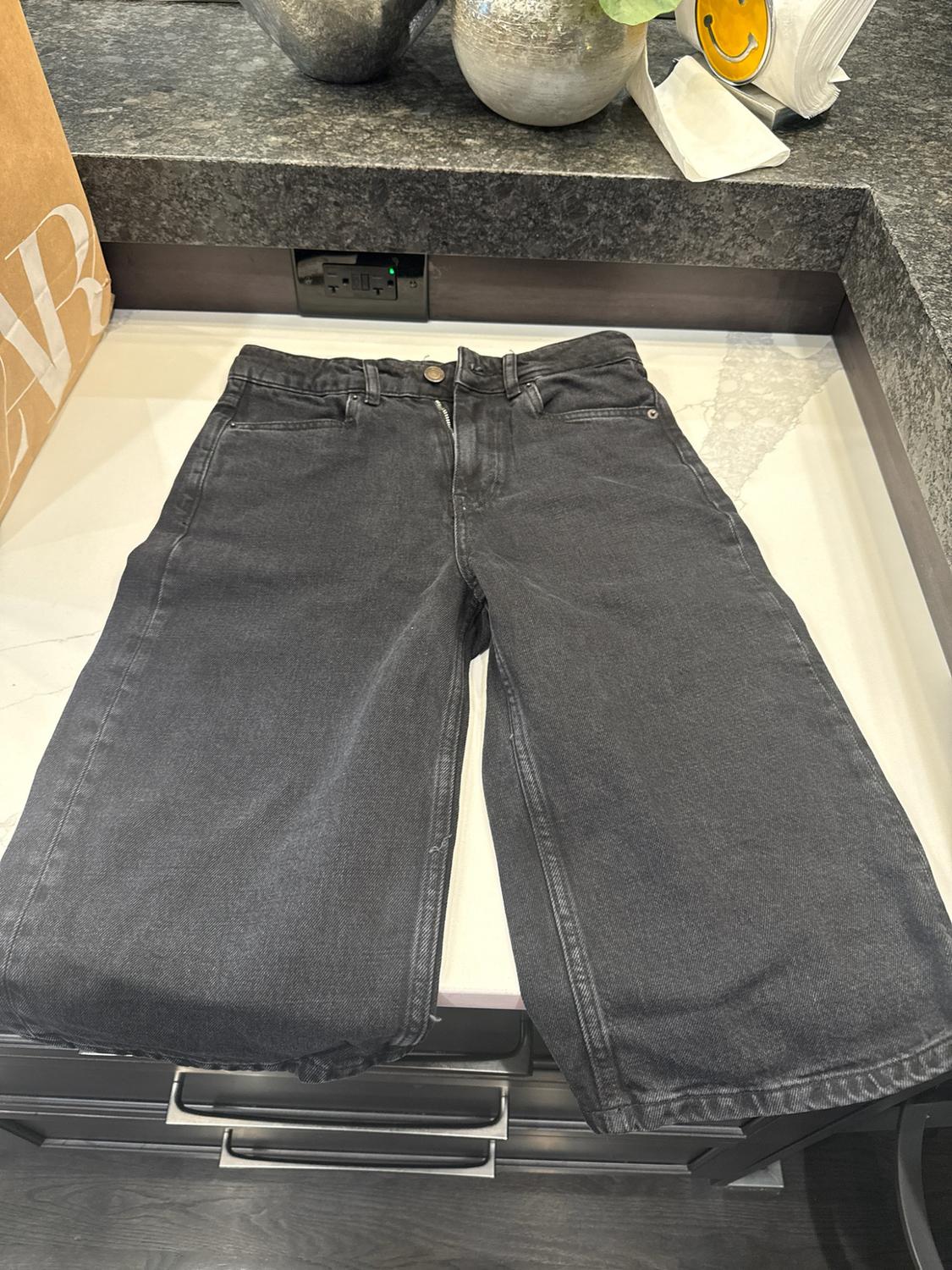 Black denim jeans on a white countertop. In the background, a paper bag and decorative objects can be seen on dark granite.