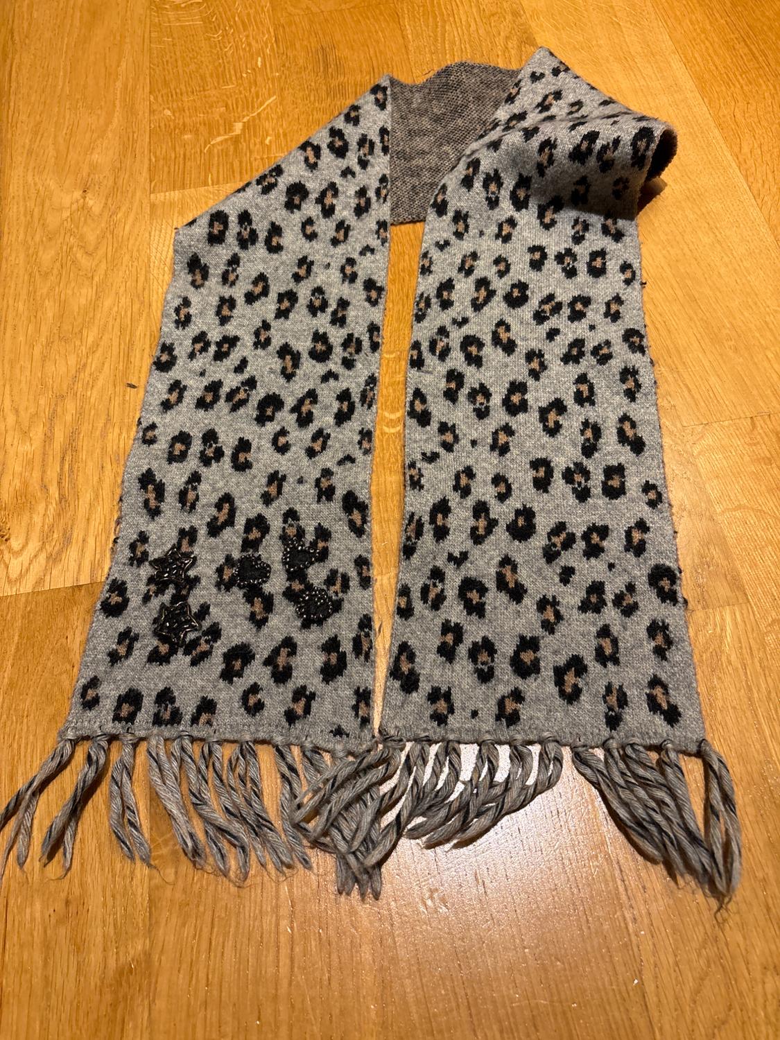 Grey leopard-print fringed scarf, decorated with dark embellishments, on a wooden floor.