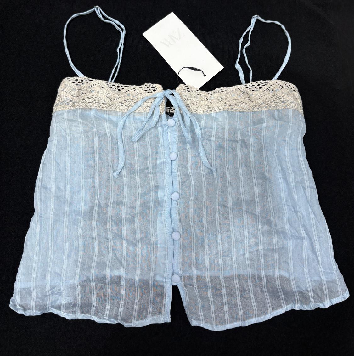 Light blue camisole with front buttons and lace trim on the neckline, on a black background. White label with "NWZ" logo.