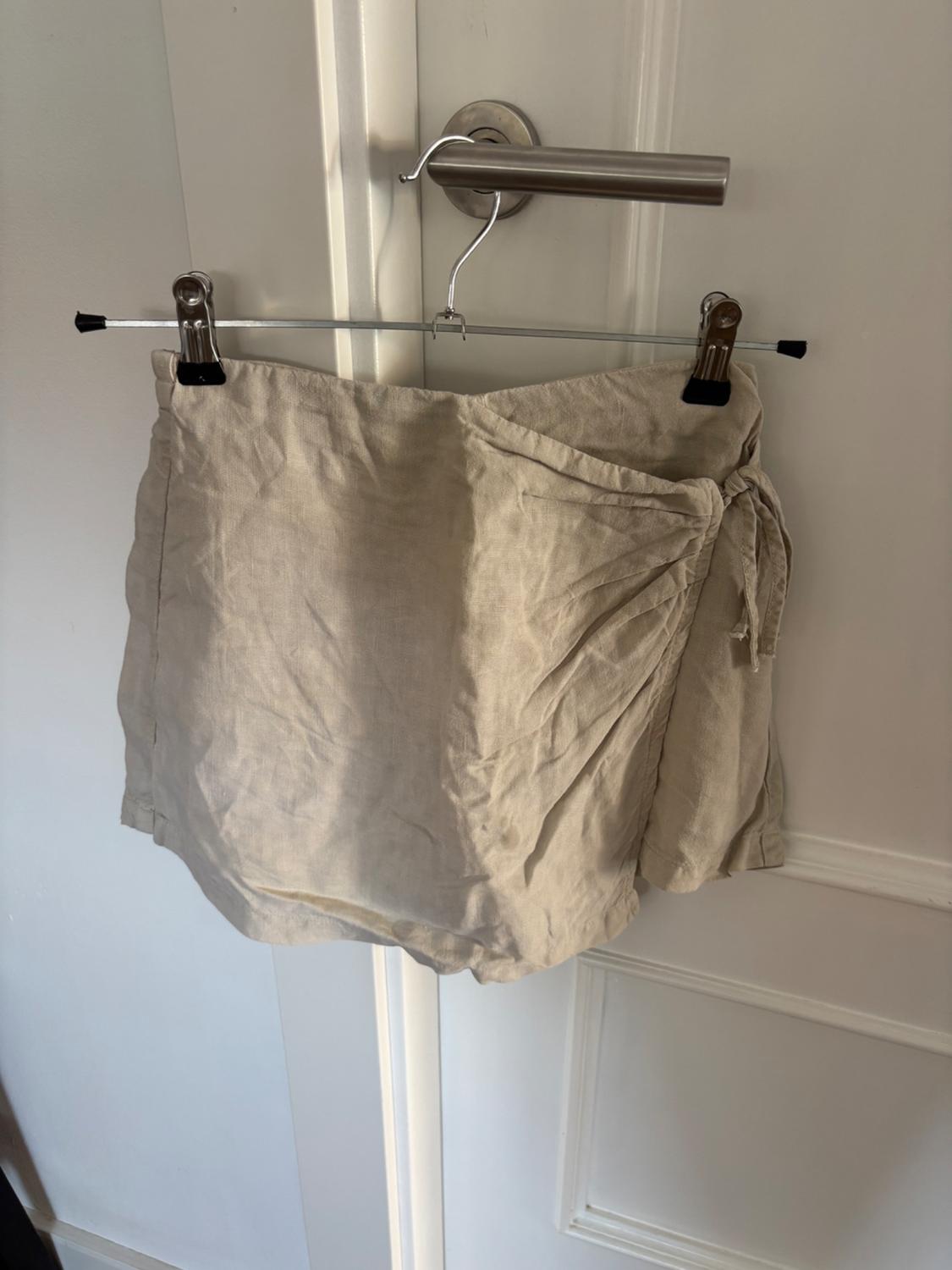 Beige mini skirt hung on a metal hanger on a white door knob. Wrinkled fabric with side tie.