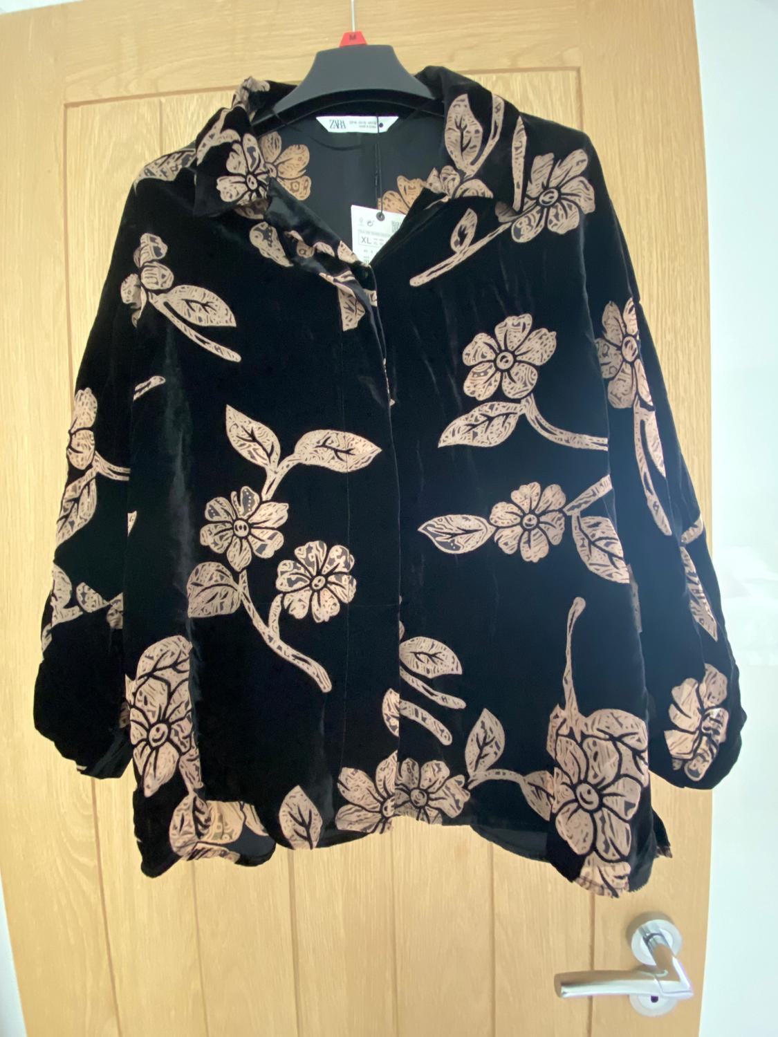 Black velvet jacket with a beige floral pattern, hanging on a hanger in front of a light wooden door.