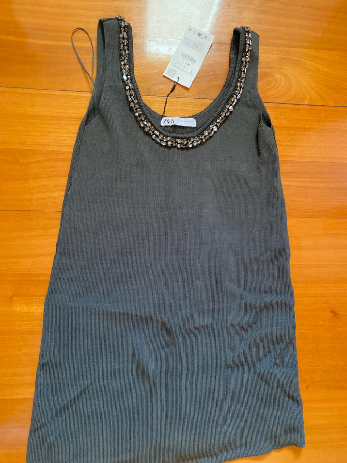Grey sleeveless top with a sparkling beaded neckline, visible label with "ZARA" text, on a wooden background.