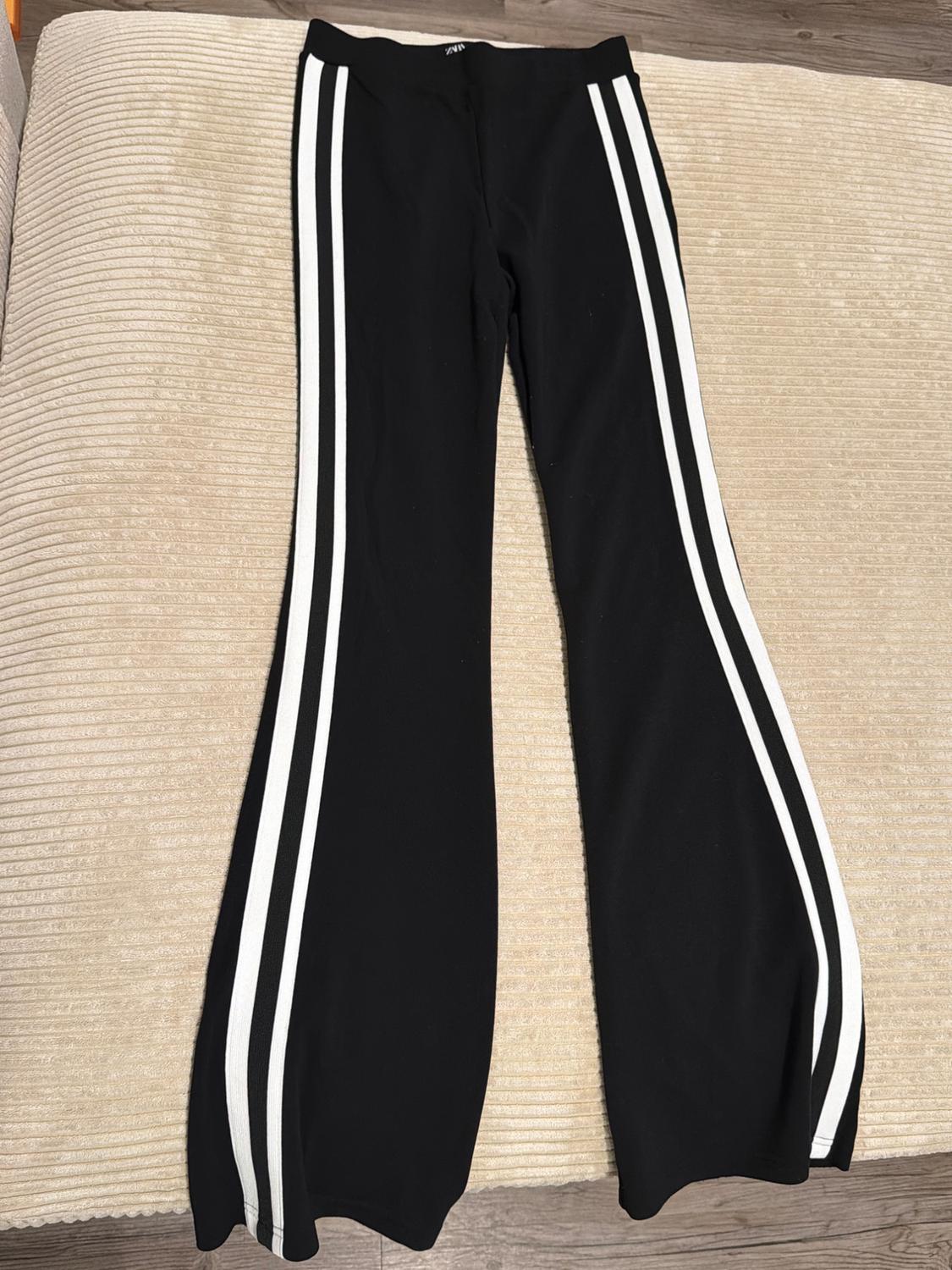 Black flared pants with two white side stripes, laid out on a textured beige surface.