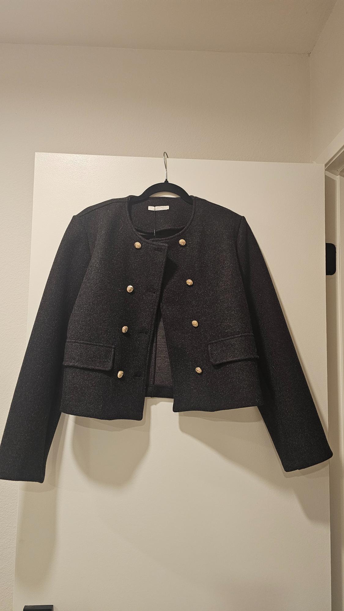 Black wool jacket with gold buttons hanging on a hanger over a white door in a minimalist interior setting.
