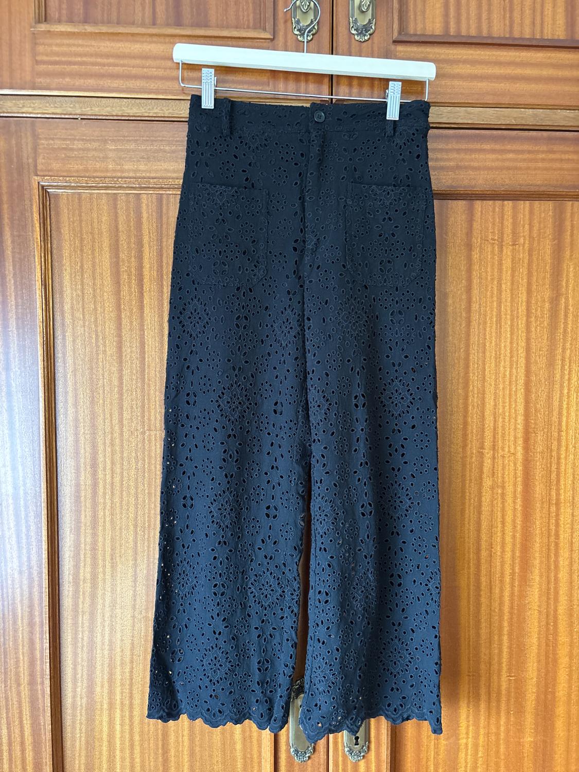 Black lace trousers hanging on a white hanger in front of a wooden door. Floral design with front pockets.