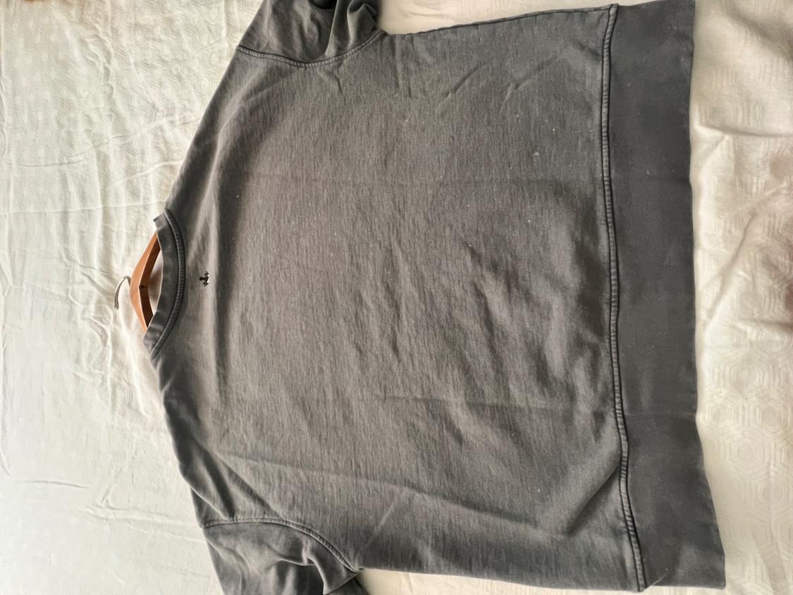 Gray sweatshirt on a white bed, with a small black logo on the chest. The garment is laid out flat and has a casual style.