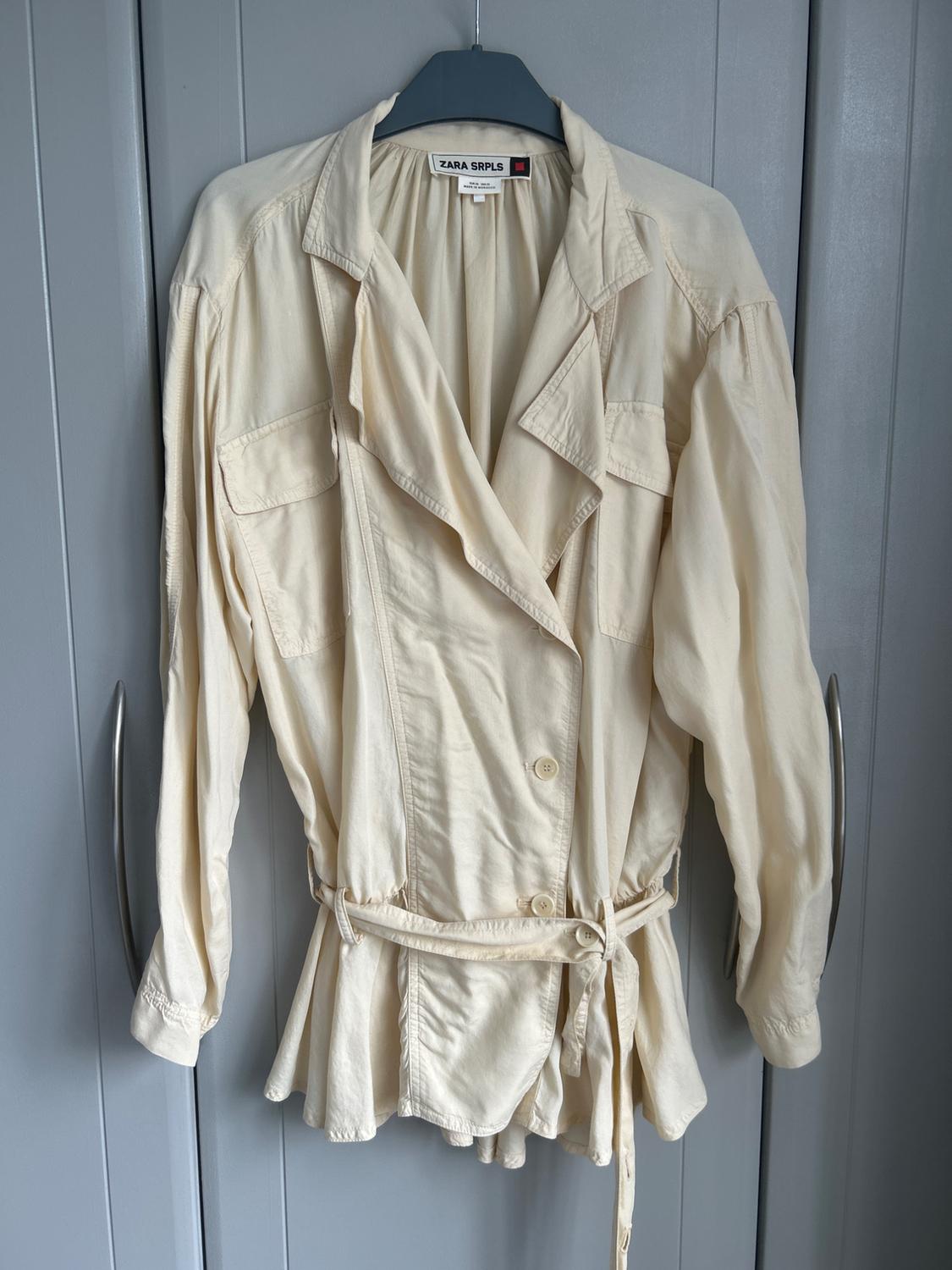 Lightweight beige jacket with a belt and pockets, hanging in a grey wardrobe. Visible text: ZARA SRPLS. Casual and relaxed style.