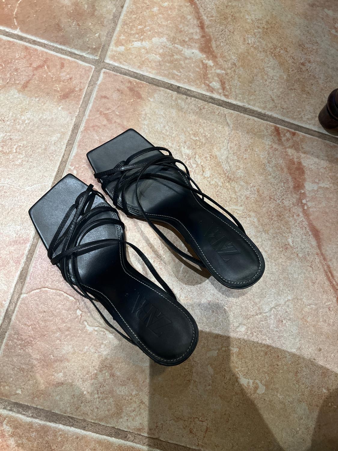 Black thin-strapped square-toed sandals on a brown tiled floor. Visible text: "ZARA".