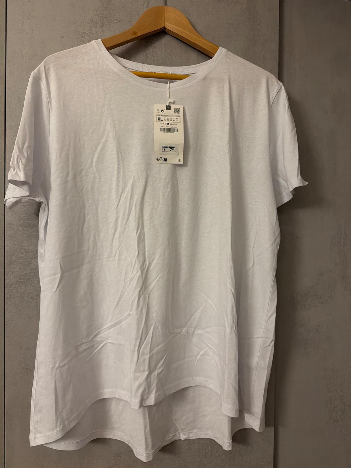White short-sleeved T-shirt hanging on a wooden hanger, with a visible label indicating size XL. Grey background.
