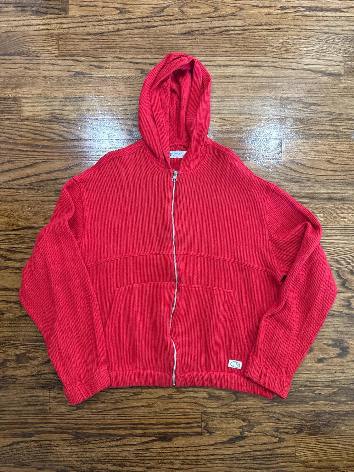 Red knit hoodie with a hood and a front zipper, laid out on a wooden floor. The interior ZARA label is visible.