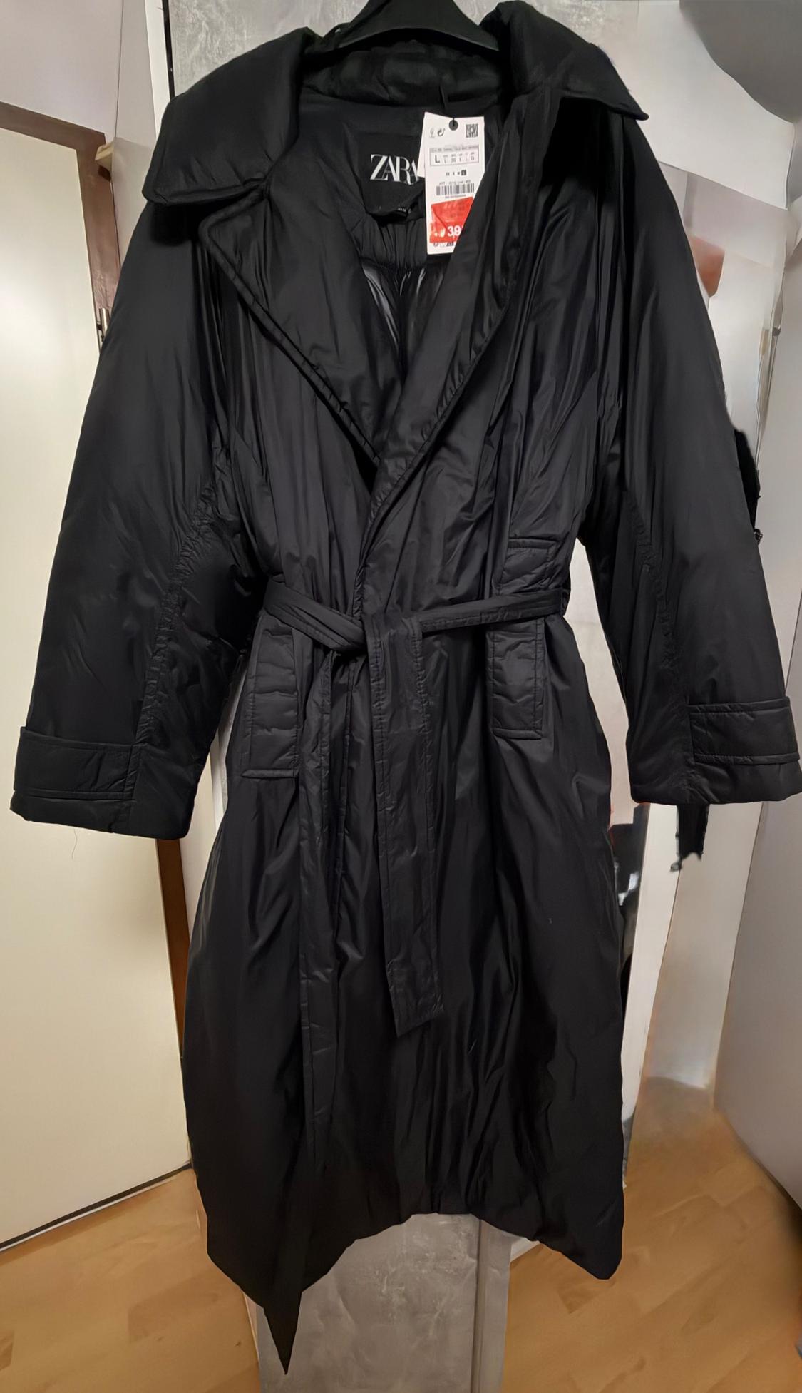 Long black coat with belt hanging on a hanger. Visible tag with a price of 39 euros. Interior room background.