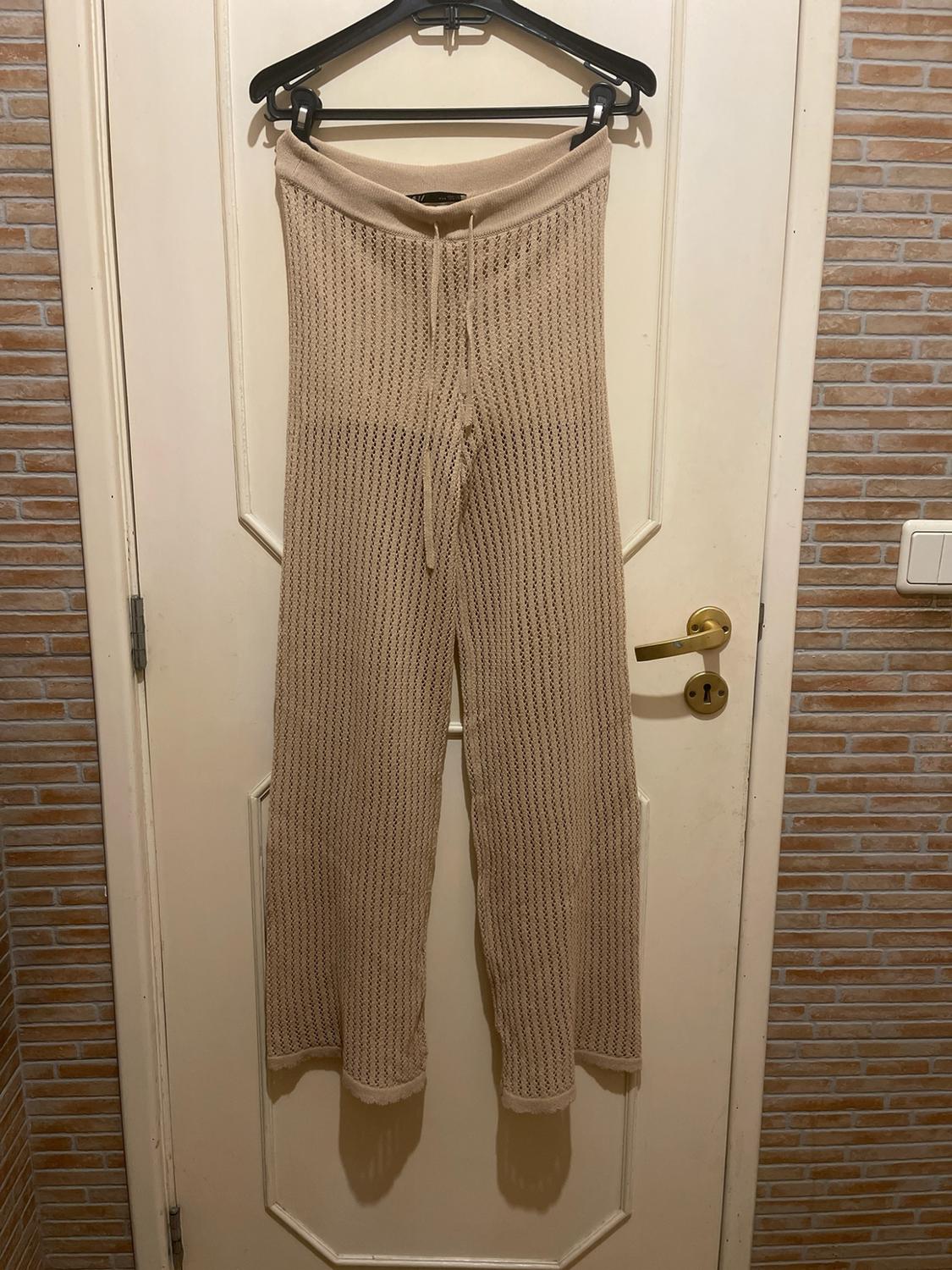 Beige open-knit trousers with a drawstring waist, hanging from a black hanger on a white door against a brick wall background.
