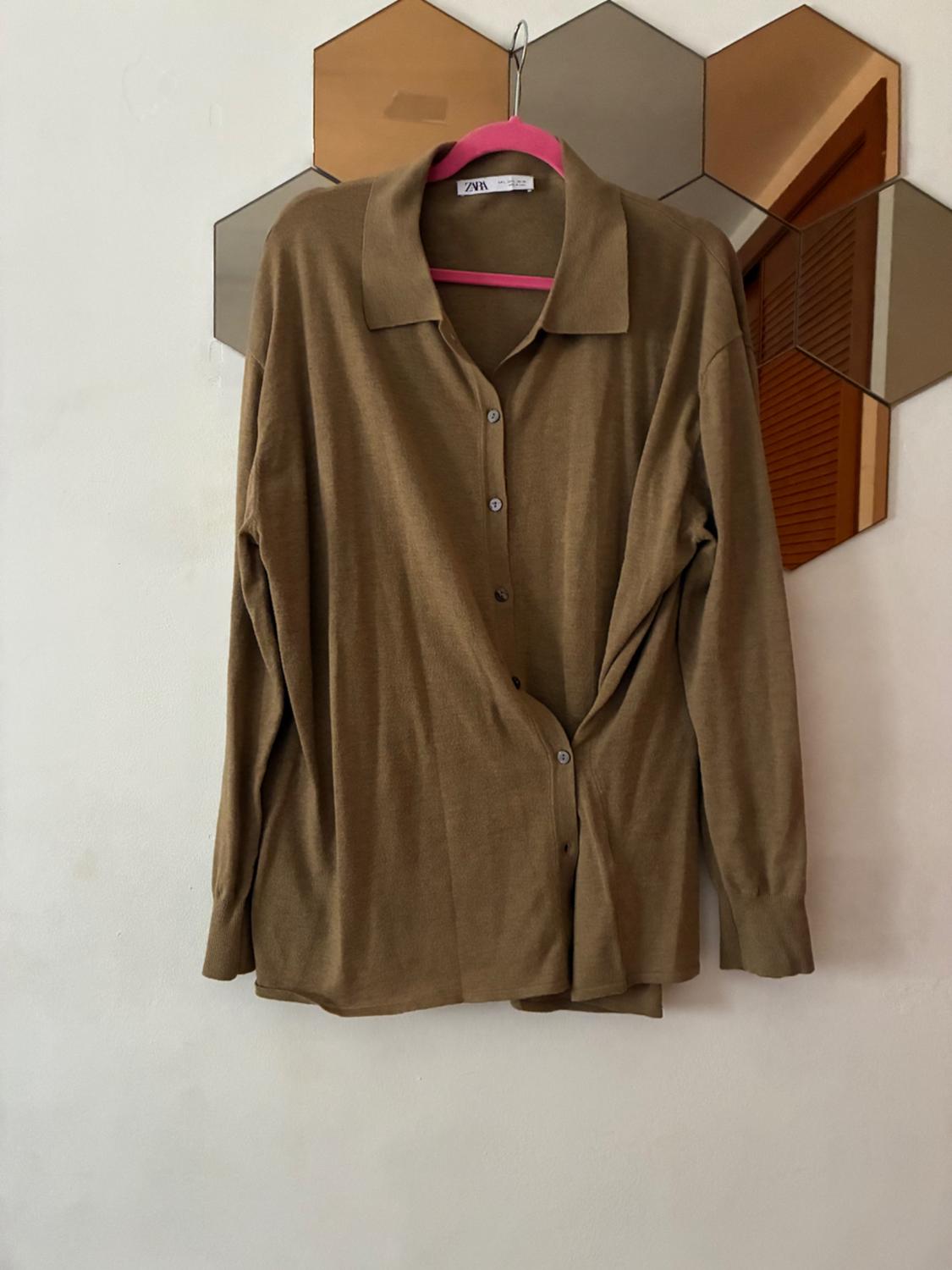 Camisa marrón de manga larga con botones colgada en una percha rosa frente a un espejo hexagonal en una pared blanca. Texto: ZARA.