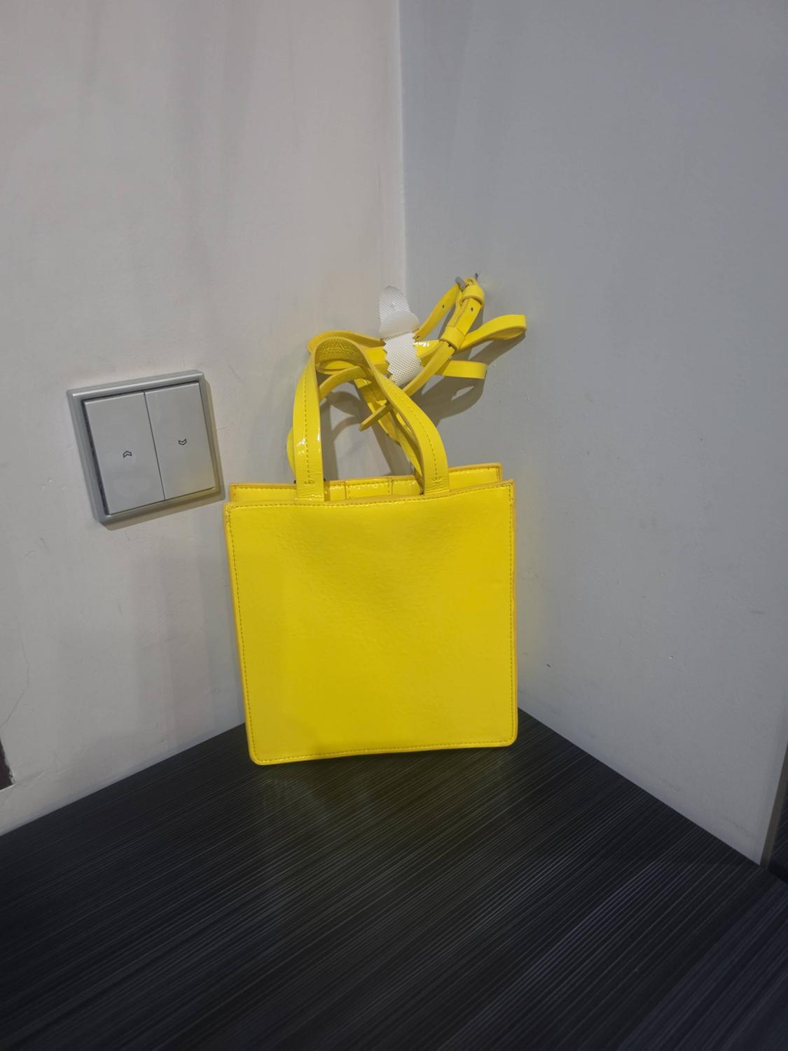 Bright yellow handbag with handles and strap tucked in, on a dark surface in the corner of white walls next to a light switch.