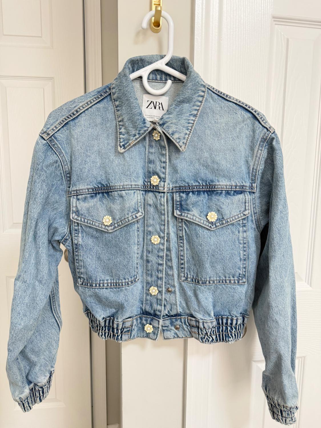 Blue denim jacket with flower buttons, elastic cuffs and hem, hanging on a white door. Tag with the text ZARA.