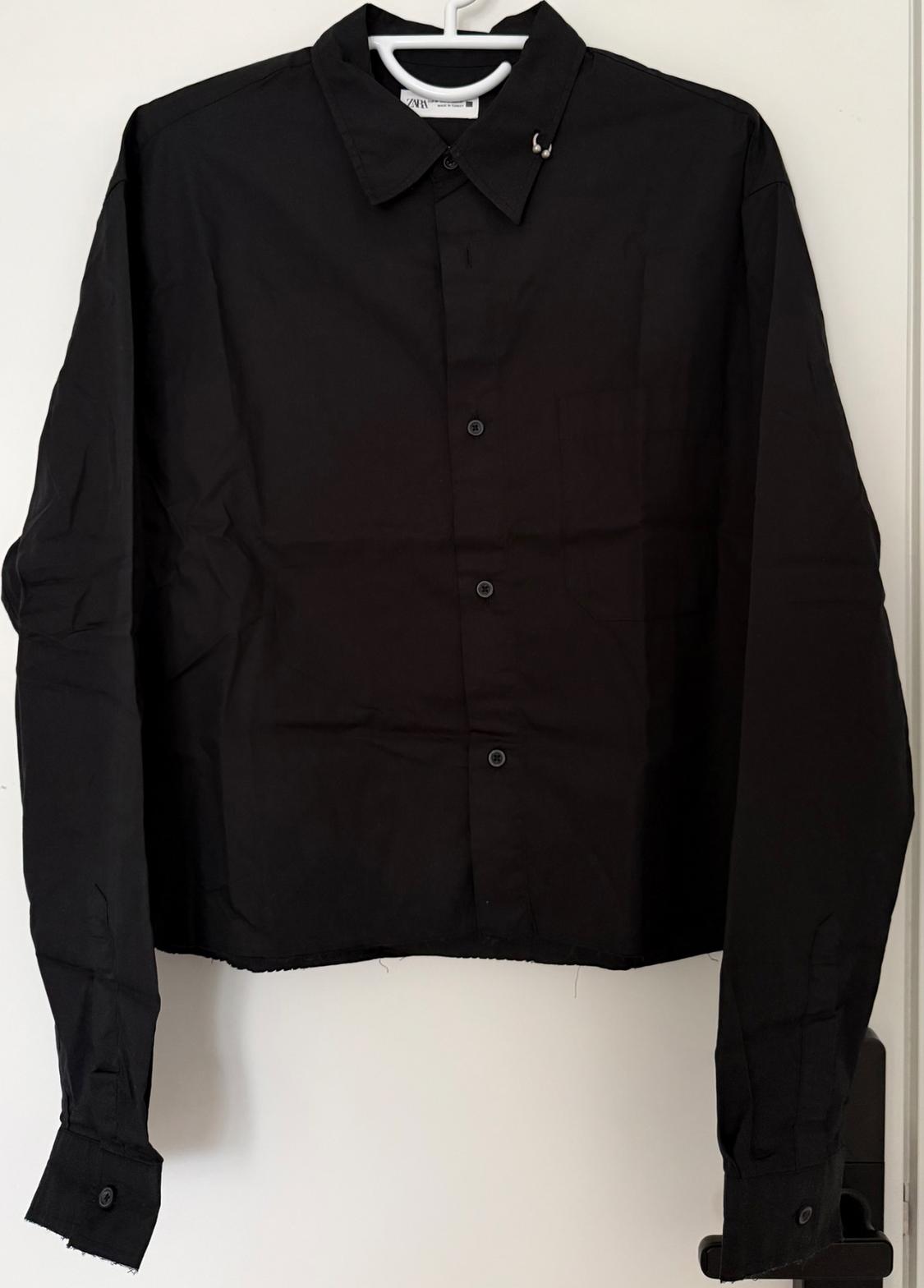 Black long-sleeved shirt hanging on a white hanger against a light background. Simple design with front buttons.