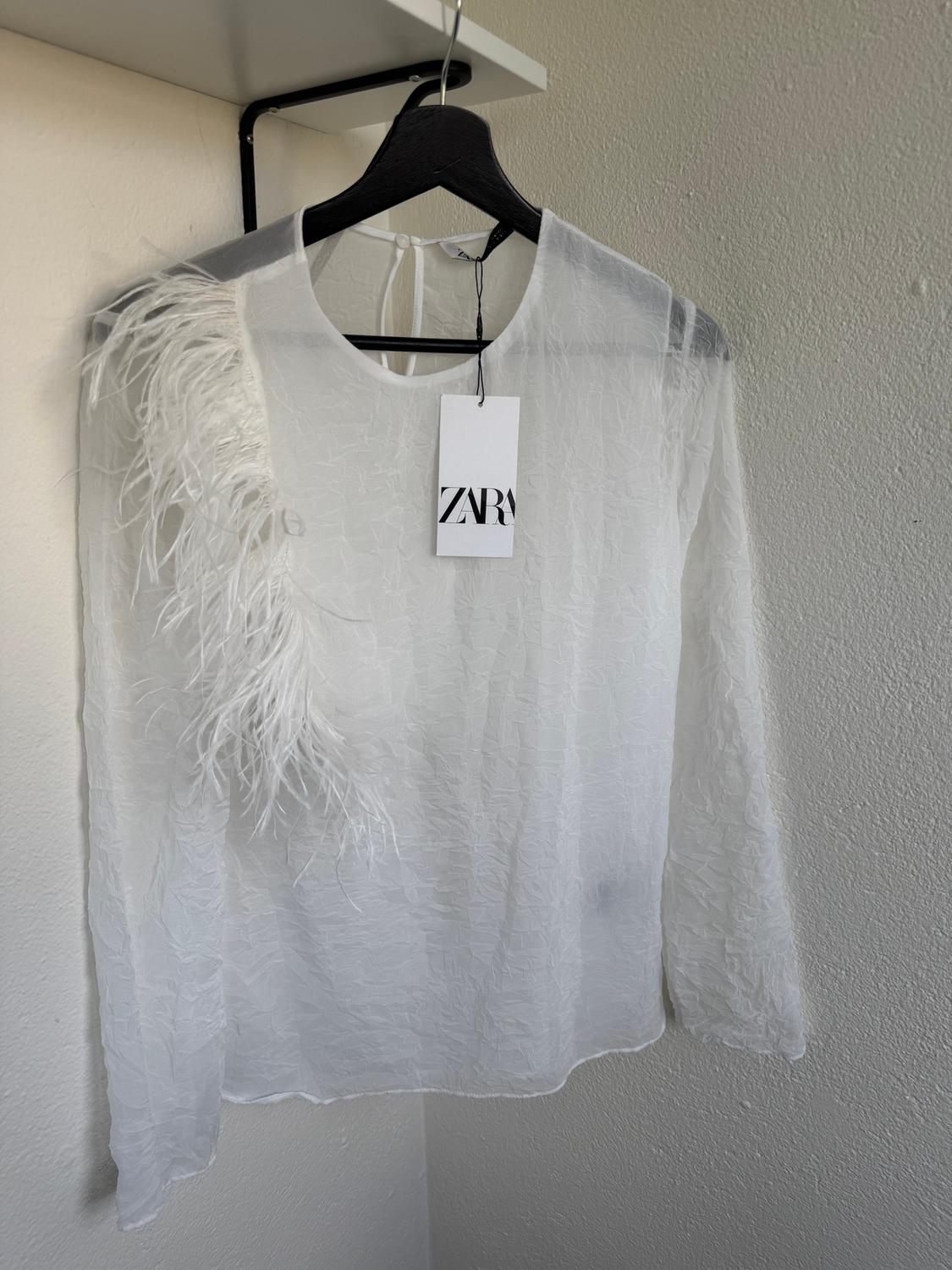 Semi-sheer white blouse with feathers on the left shoulder, hanging on a black hanger. Label with text "ZARA".
