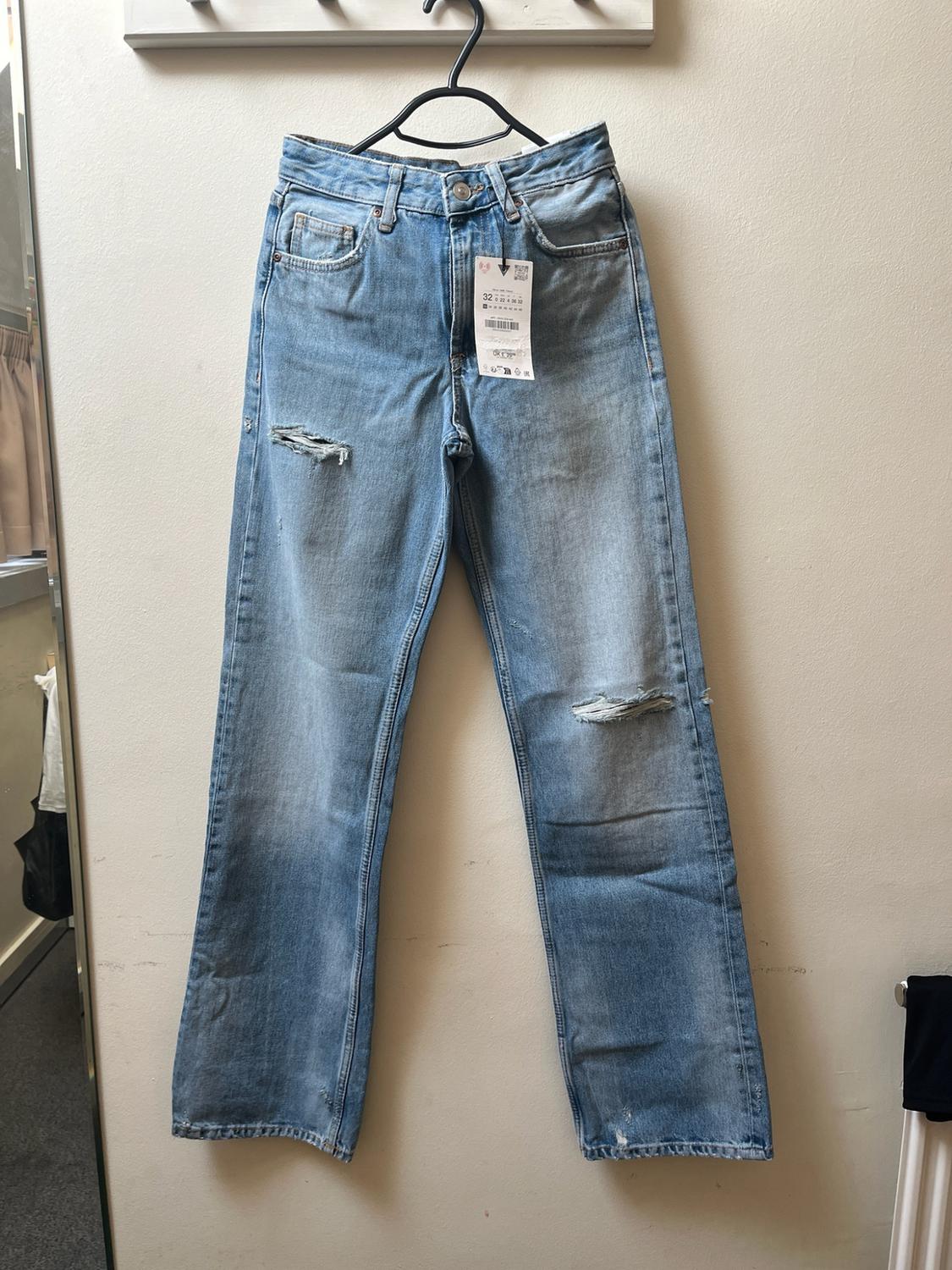 Light blue ripped jeans hanging from a black hanger on a light-coloured wall. A label with the number 32 is visible. Relaxed atmosphere.