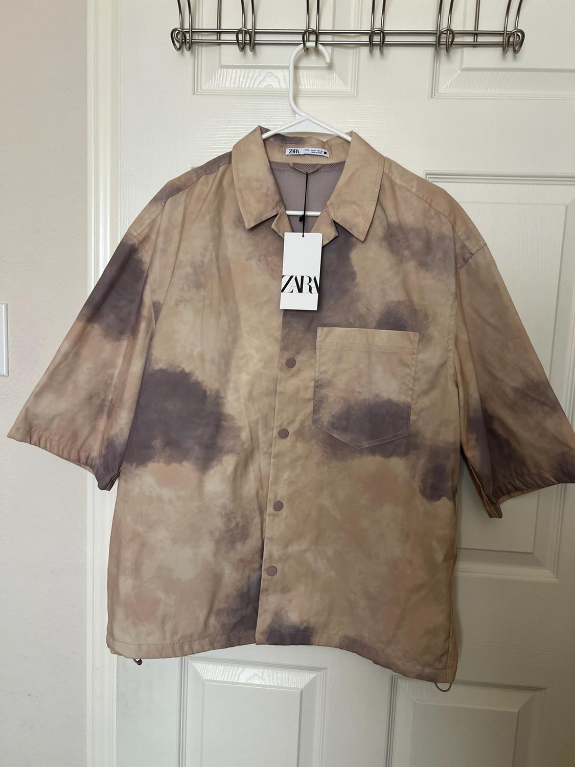 Beige short-sleeve shirt with a grey faded print, buttons, and front pocket, hanging on a door. Label with the text ZARA.