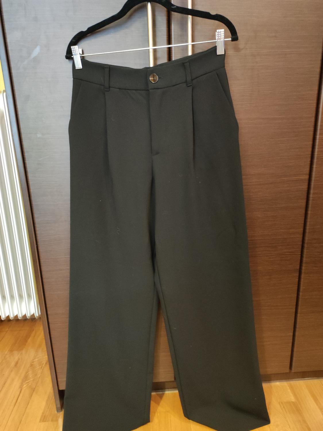 Black dress trousers hanging on a black hanger in front of a wooden wardrobe. Wooden floor visible.
